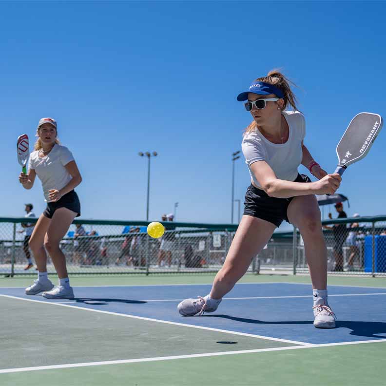 Two women wearing white shirts and black shorts playing pickleball outside on a court App Partnership page quote image