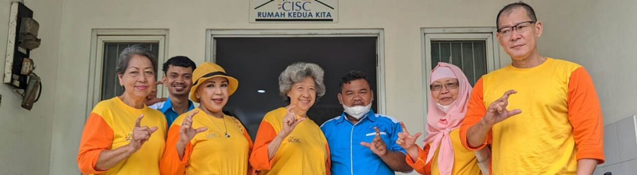A group of eight people stand in front of a building entrance beneath a sign that reads “CISC Rumah Kedua Kita.” Six wear matching yellow‑orange shirts, one wears a blue shirt, and one wears a pink hijab. They face the camera and make a “C” hand gesture, appearing to pose as a unified group