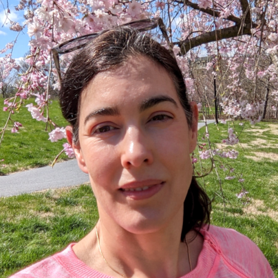 Woman, dark brown hair in a ponytail, dark framed eyeglasses on top of head, pink sweater, pink flowers in background