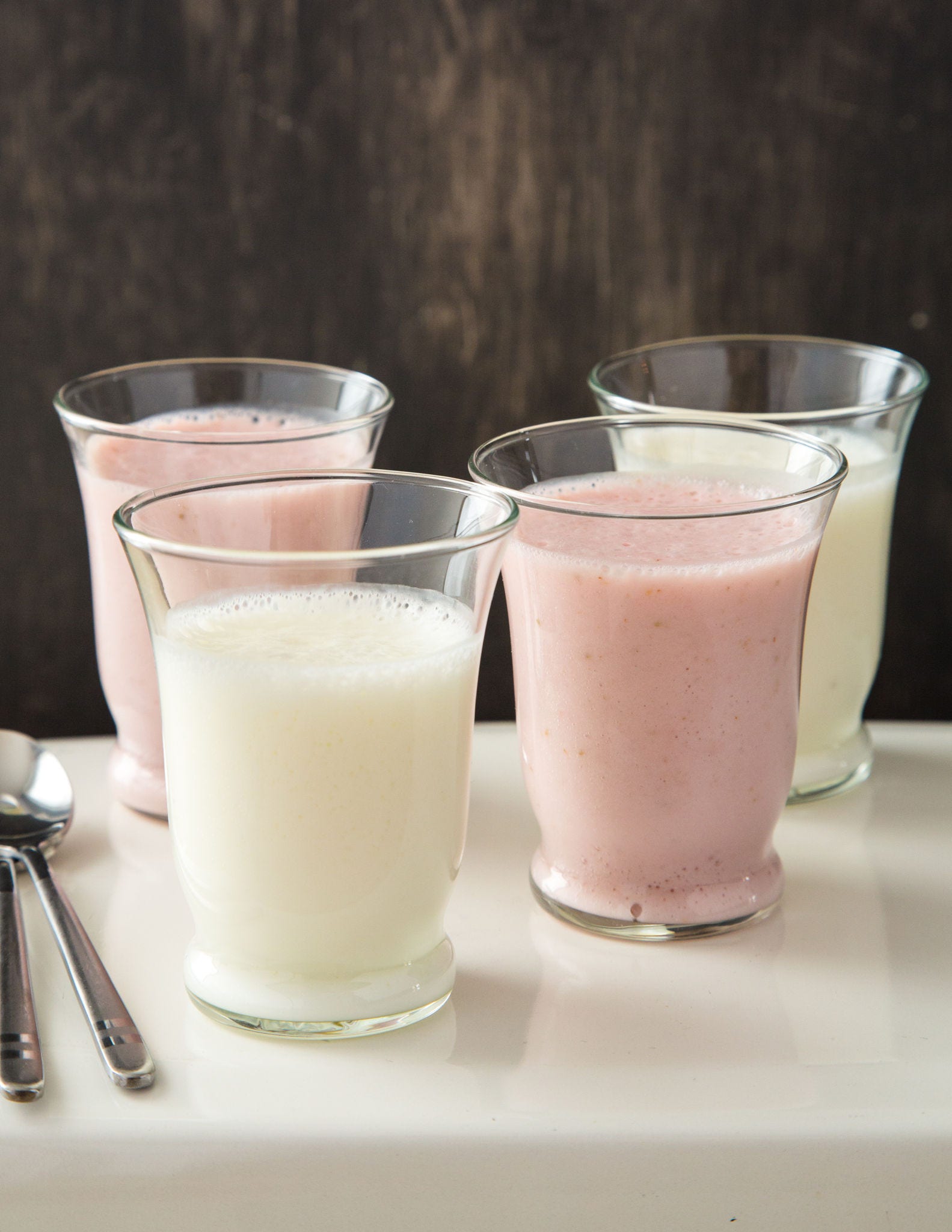 Two glasses of white pudding sit next to two glasses of pink strawberry pudding on a table in front of a wood backdrop.