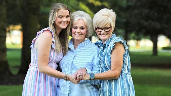 Three women standing together outdoors, smiling and holding hands.