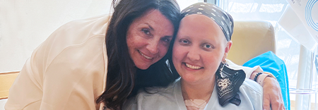 Dos personas sonriendo juntas en un entorno médico, una con un pañuelo en la cabeza y la otra con el pelo largo y oscuro, sentadas cerca de un equipo hospitalario.