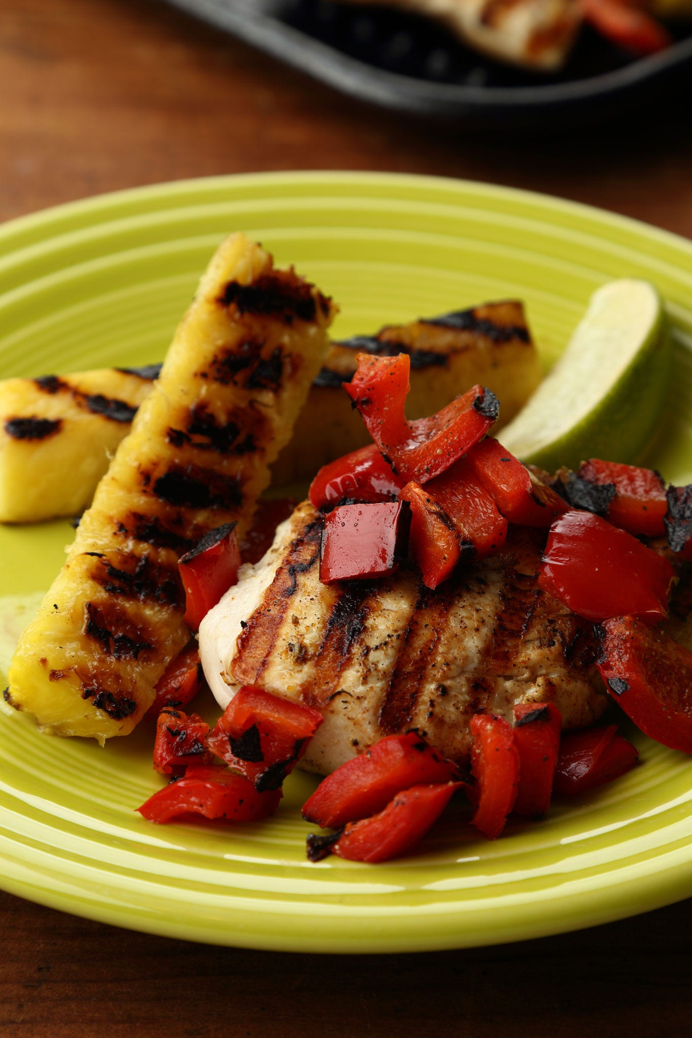 Pieces of grilled pineapple sit on a plate next to chicken topped with chopped red pepper.