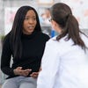 A woman wearing a black shirt and sitting on an exam table speaks to a doctor wearing a white lab coat. 