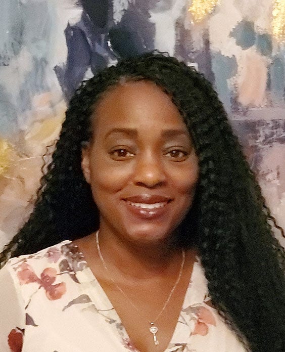 Black woman with long wavy hair, wearing flowered top and key necklace