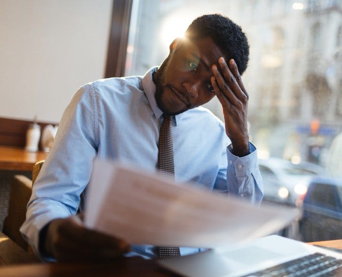 a stressed out man rubs his brow as he looks at papers while sitting at a computer