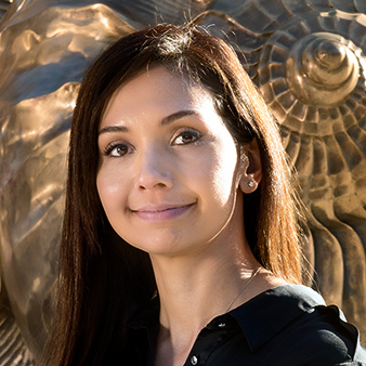 Woman, smiling, black blouse-buttons open at the top, long brown straight hair, sculpture in background