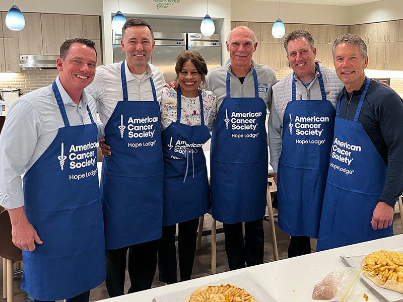 Six people wearing aprons posing in kitchen, smiling together.