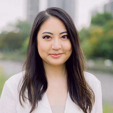 woman with brown hair flowing on both shoulders, white jacket in front of blurred cityscape