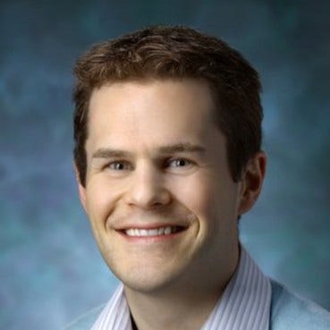 man with brown curly hair, wearing blue & white striped shirt under light blue sweater in front of blue background