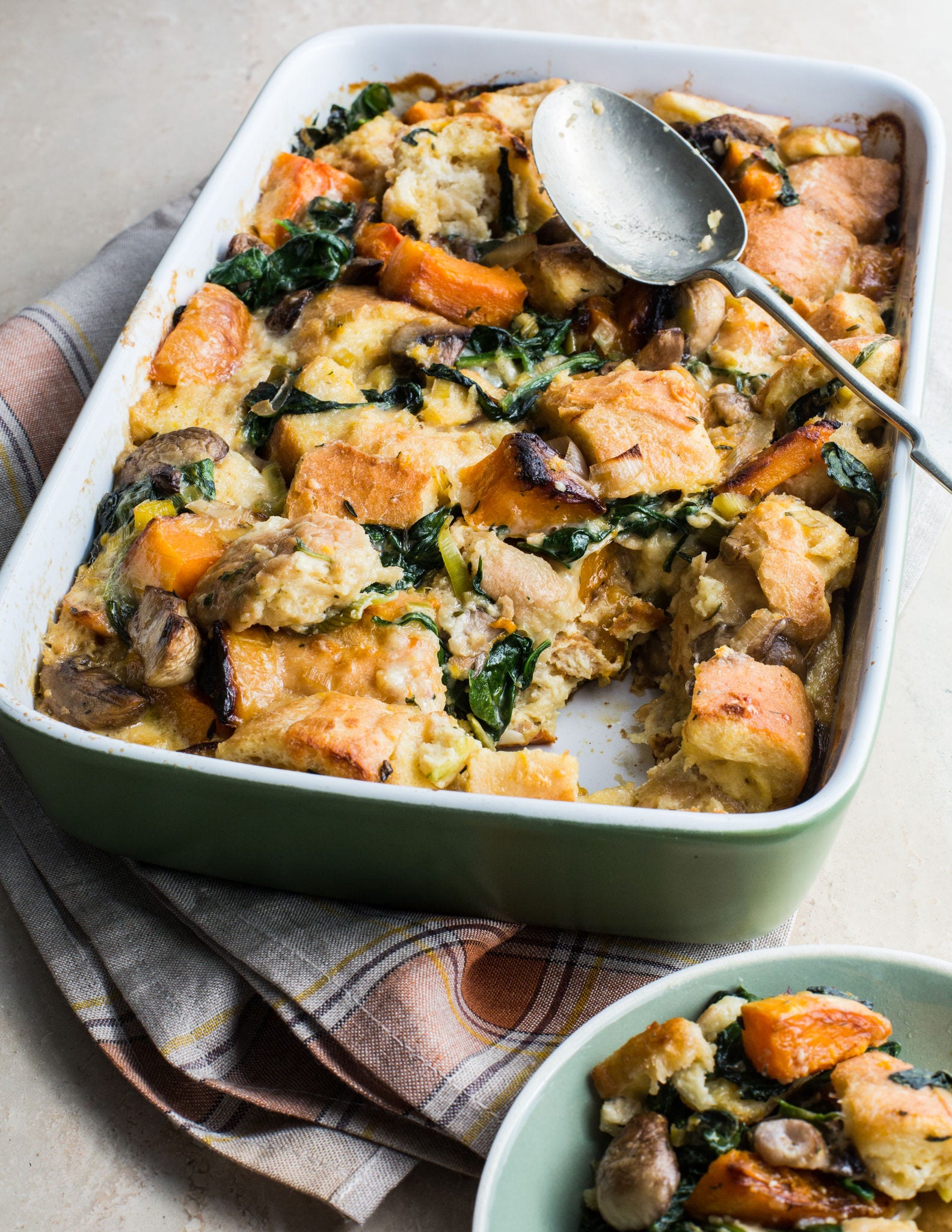 A serving spoon rests on a rectangular baking dish that holds bread pudding made with butternut squash, mushrooms, and spinach.