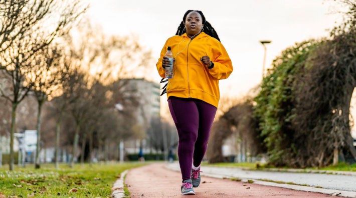A woman wearing a yellow sweatshirt and maroon pants jogs along a paved path with trees and buildings in the background.