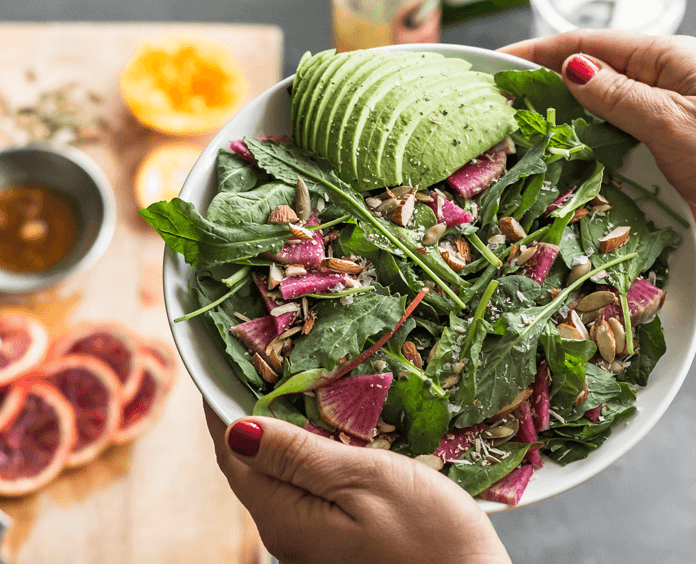 Woman's hands holding a bowl of salad with other foods on a counter behind