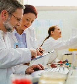 A male and female researcher examine information together in a lab setting with another female researcher in the background of the picture.
