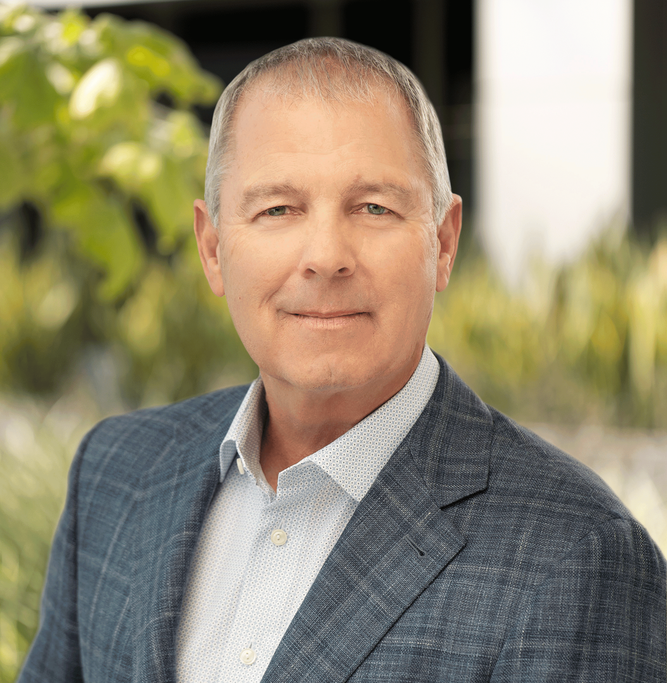 Man in suit jacket posing outdoors with greenery behind.