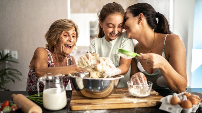 A woman presses her face against a girl standing next to an older woman as they all mix dough in a bowl with their hands. 