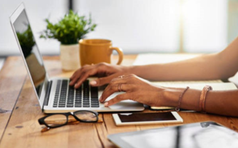 wooden desk with glasses, mug, plant and hands typing on laptop