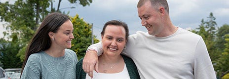 Three people standing outdoors with trees and cloudy sky in the background, smiling and leaning together.