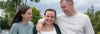 Three people standing outdoors with trees and cloudy sky in the background, smiling and leaning together.