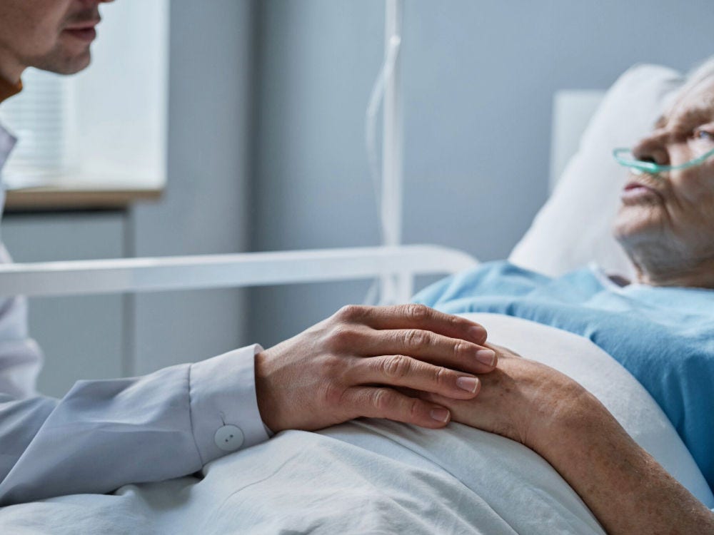 Image of a younger man comforting and talking to an older person who is lying in a hospital bed. The younger man is resting his hand on the older person's folded hands.