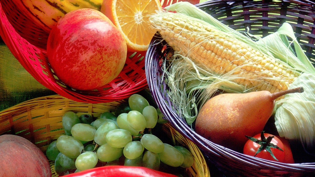 three different colored woven baskets in a tight frame containing various fruits and vegetables such as grapes, a peach, an orange, ear of corn, red pepper, tomato and a pear