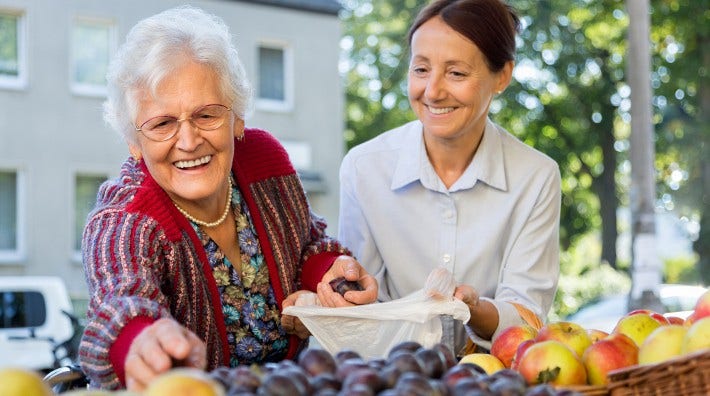 caregiver helping a senior woman bag fruit at an outside market