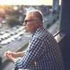 A man with gray hair stands on a balcony overlooking a street and parked cars with a city skyline visible in the distance.