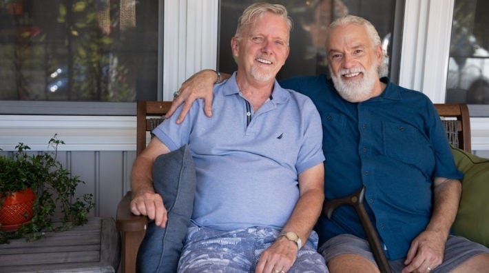 Steve Drayton and his Husband, Phen, smile while sitting on a porch bench