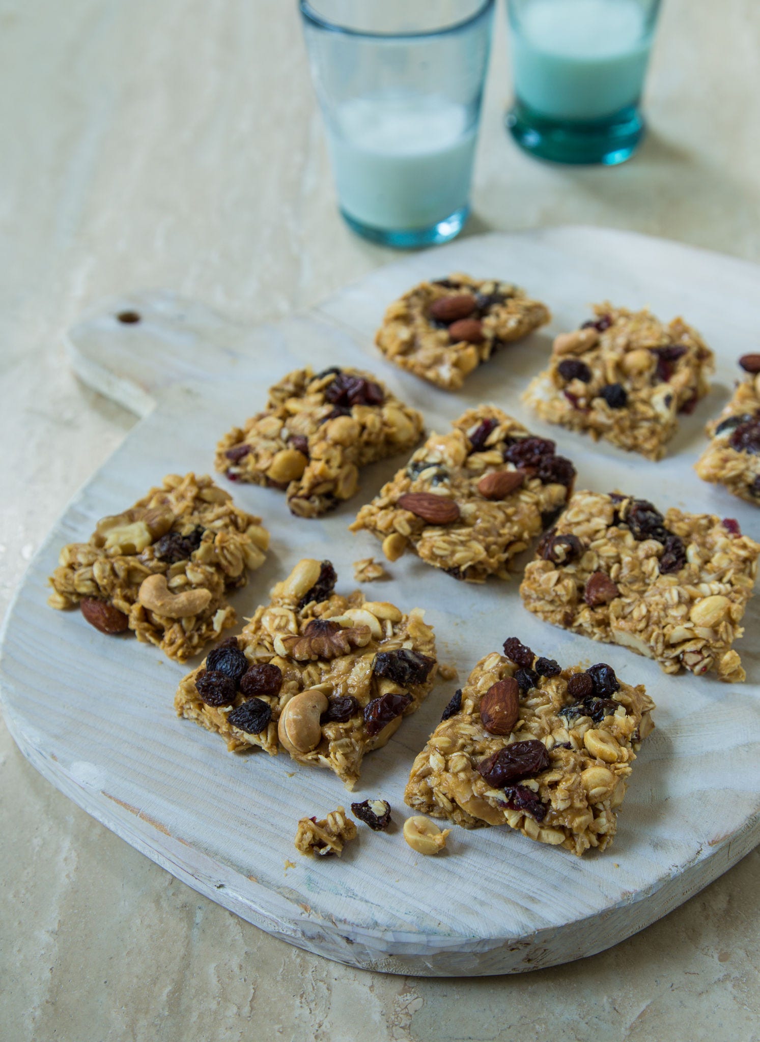 Square pieces of granola bars lay side by side on a cutting board with two glasses of milk in the background. 