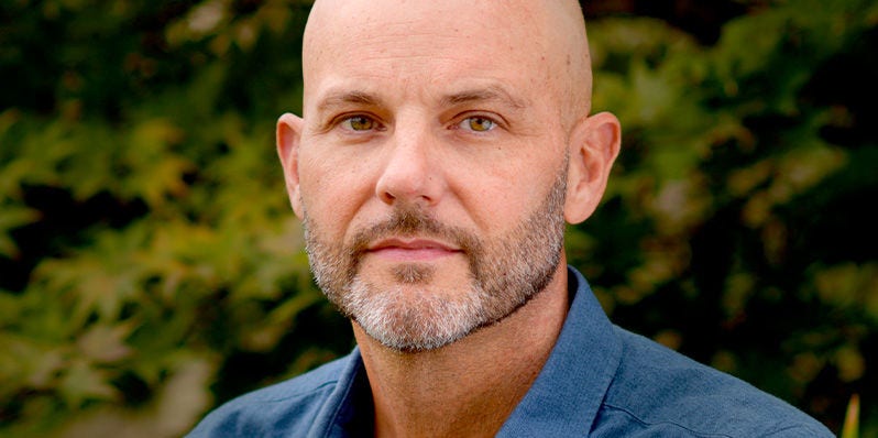 Hombre con la cabeza rapada y barba sonriendo suavemente al aire libre.