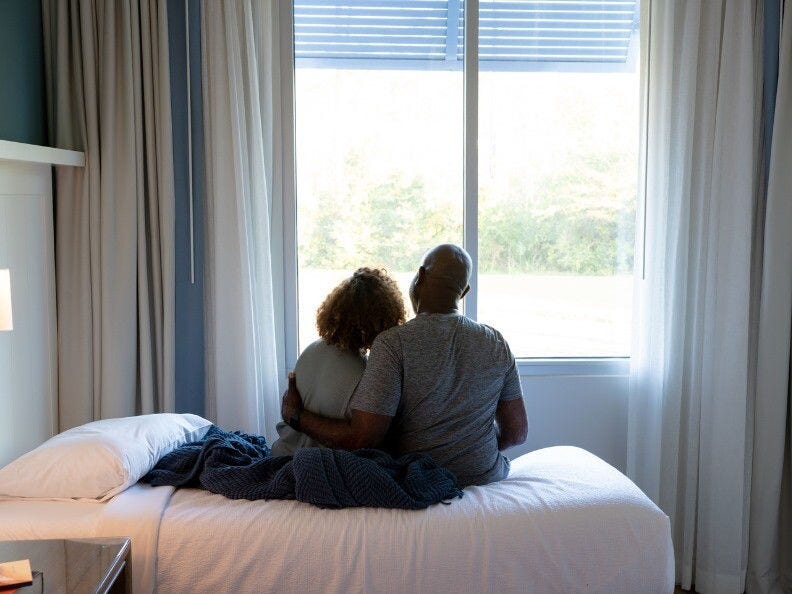 black man with arm around woman while sitting on bed looking out the window