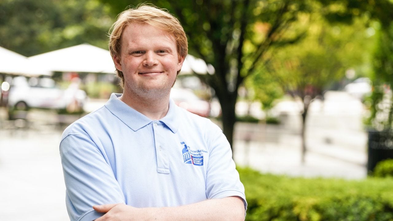 A man with red hair, wearing a blue shirt with the American Cancer Society logo, smiles while posing for a photograph