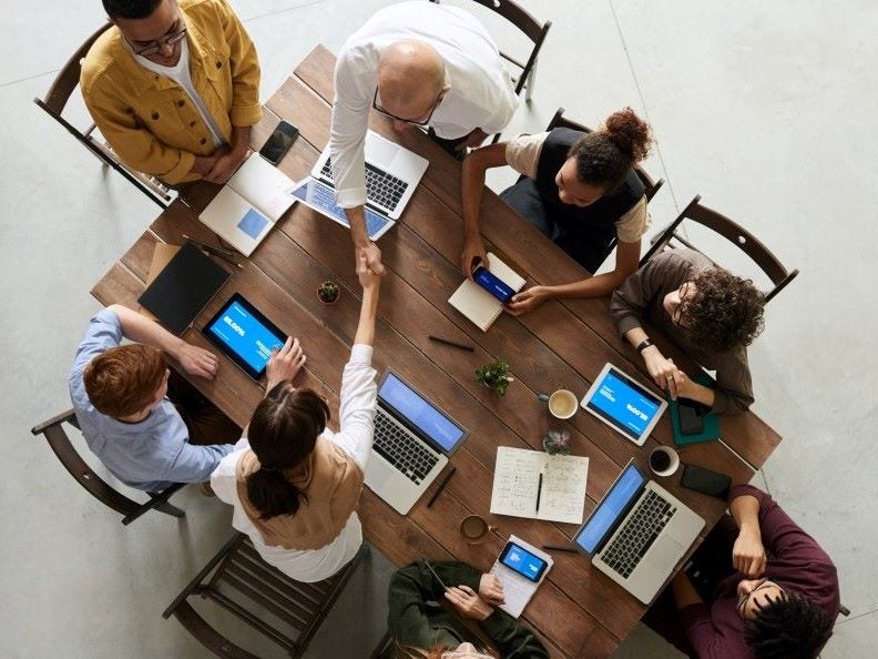 Group of people sitting around a table with devices such as laptops, tablets, cell-phones and in discussion, top down camera view