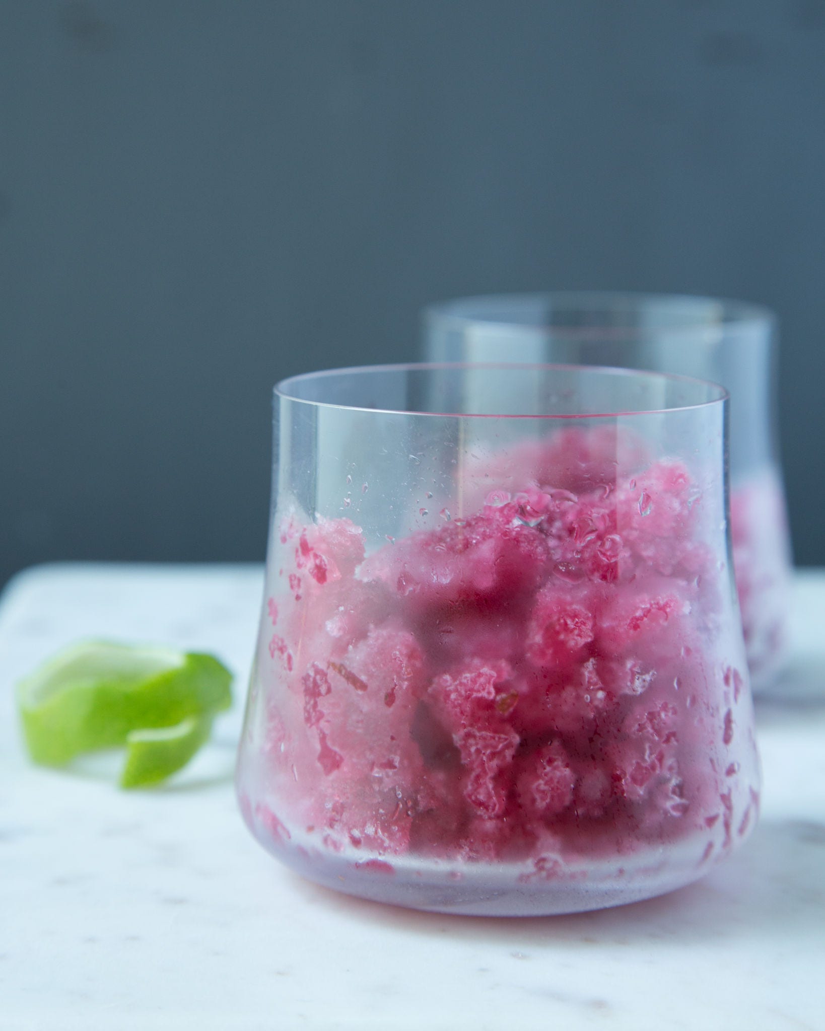 Frozen pieces of dark pink cranberry granita sit in a glass in front of a gray background.