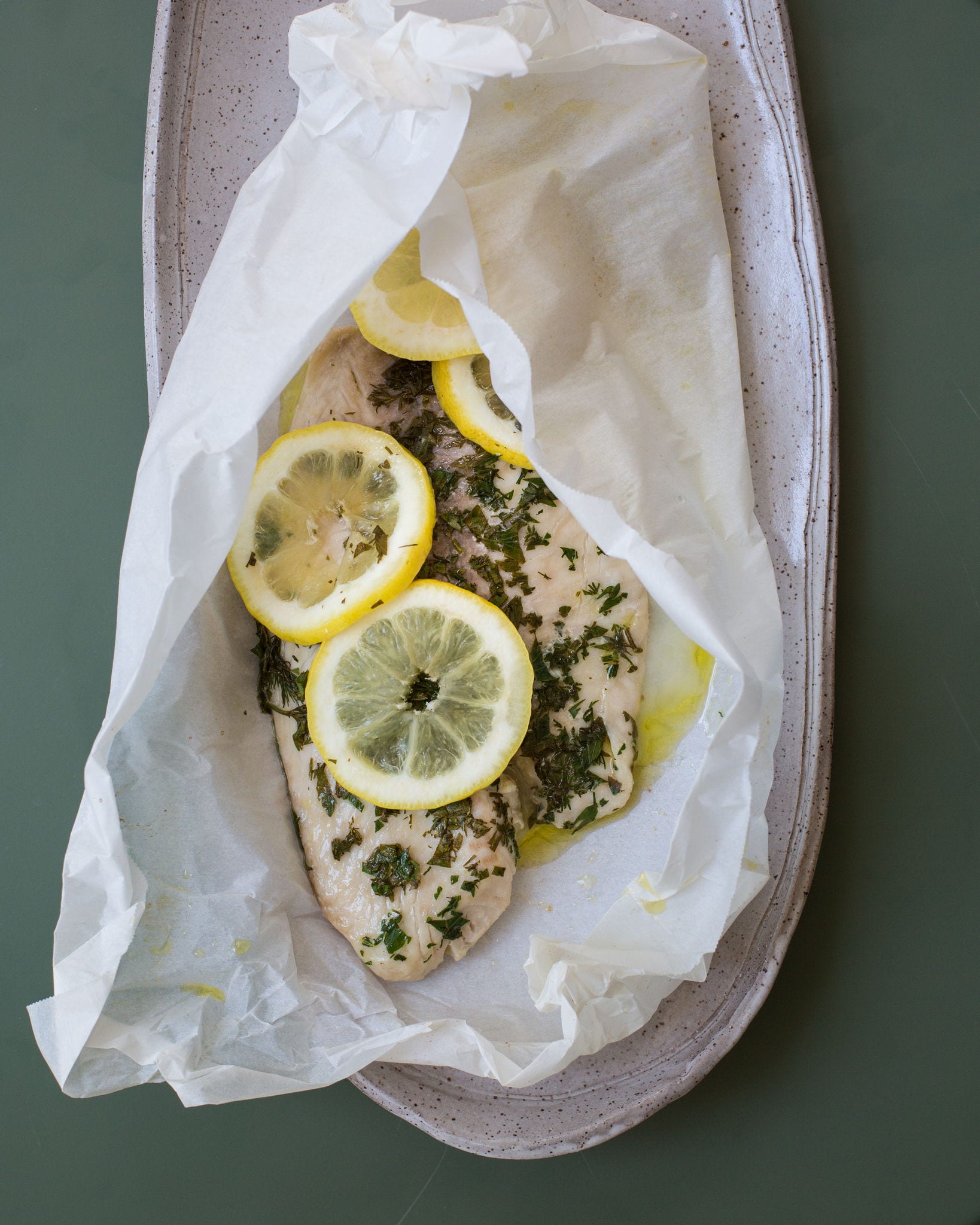 Seen from overhead, a small platter holds a piece of tilapia topped with lemon slices and wrapped in parchment paper.
