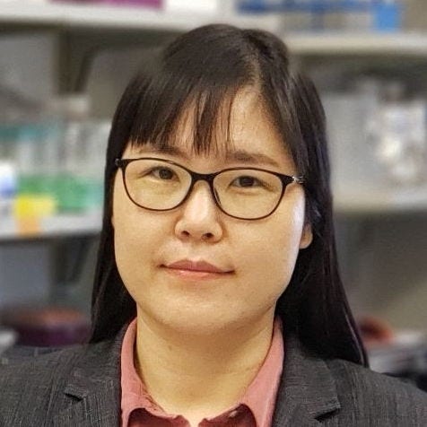 Asian woman in lab wearing gray jacket and pink shirt with bangs and glasses