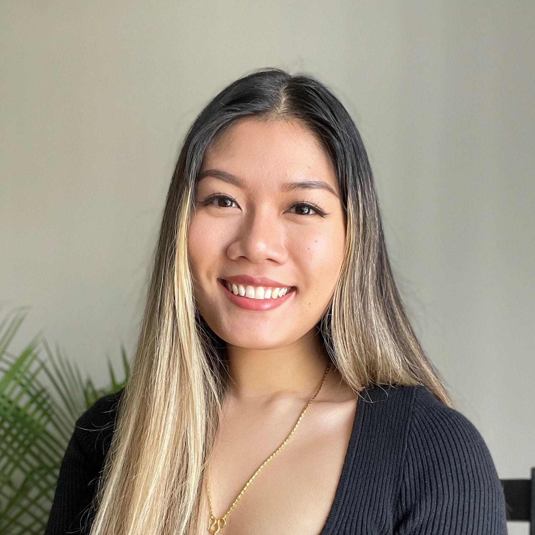 Asian woman sitting in gray chair in front of plant, blonde hair and gold necklace