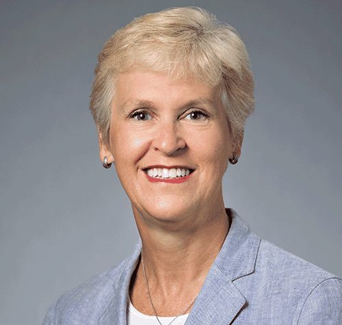 Smiling woman with short blonde hair, light blazer, gray background.