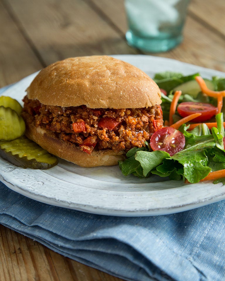 A sloppy joe made with ground turkey and quinoa sits on a white plate, with pickles on one side and greens on the other.