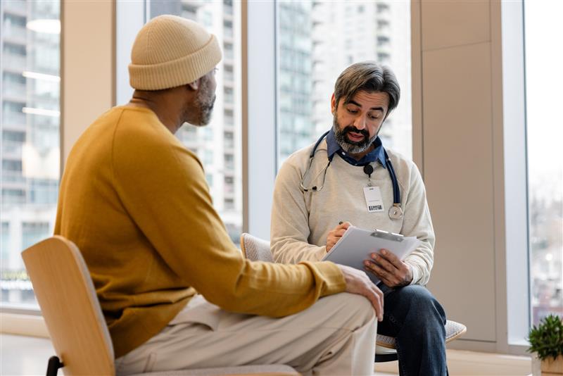 A man sits in a chair, talking to a healthcare provider with a clipboard