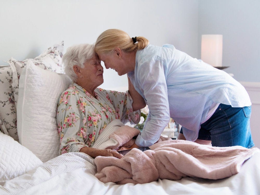Image of a middle-aged woman embracing and touching foreheads with a smiling older woman in bed