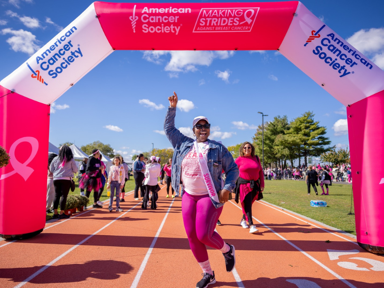 Breast cancer survivor celebrating under American Cancer Society arch.