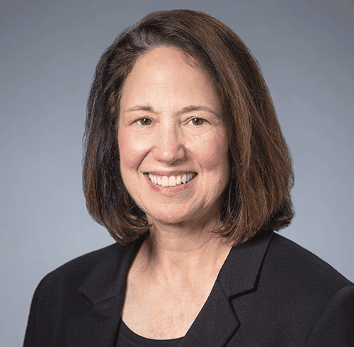 Smiling woman with shoulder-length brown hair, black blazer, gray background.