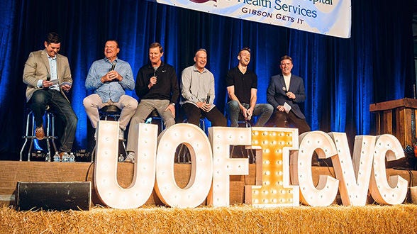 Panel of men seated on stage behind large lit letters.