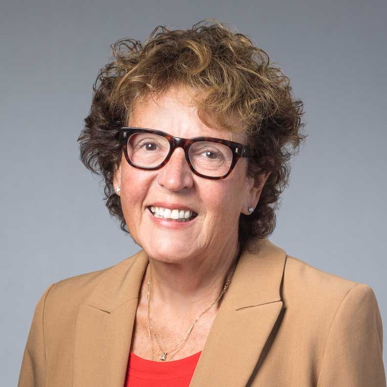 Smiling woman with glasses wearing tan blazer against gray background.