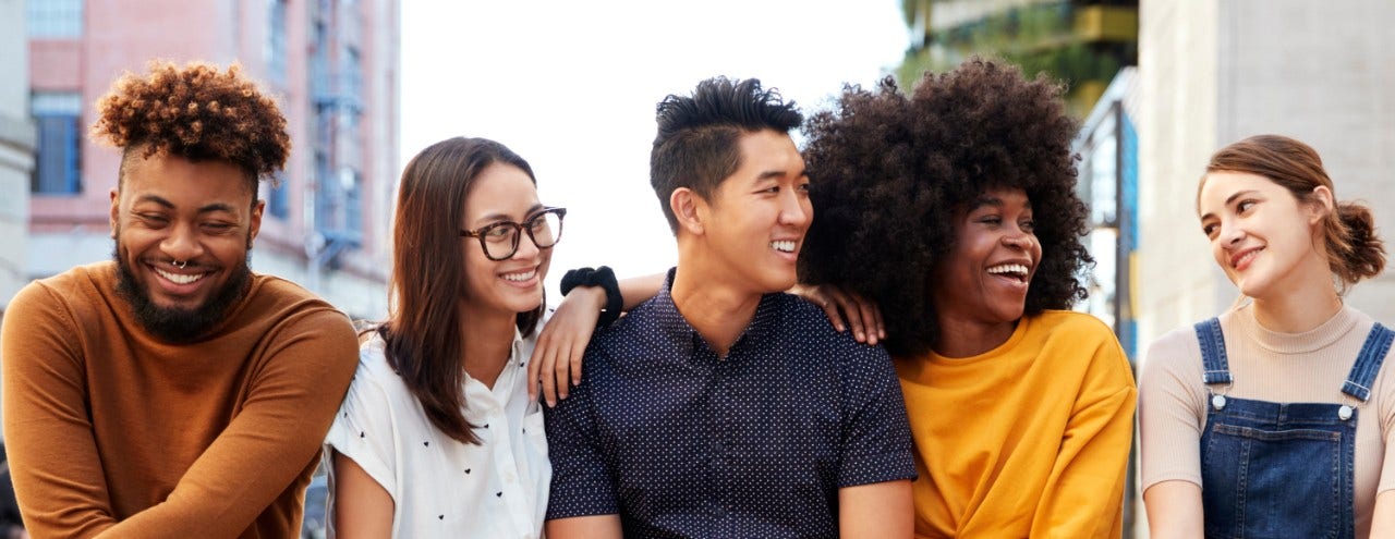 Row of college-age men and women, black man with beard and brown shirt next to white woman with long brown hair and glasses next to Asian young man next to African American woman with afro and golden yellow shirt next to white girl with pony tail in overalls