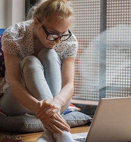 A young blonde woman with thick dark glasses is sitting on a cushion viewing her laptop.
