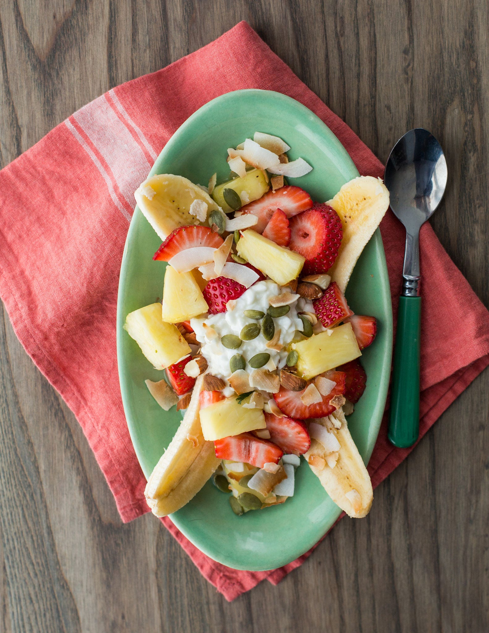 Seen from above, an oval plate holds bananas, sliced longwise and topped with strawberries, pineapple, coconut, almonds, and pepitas.