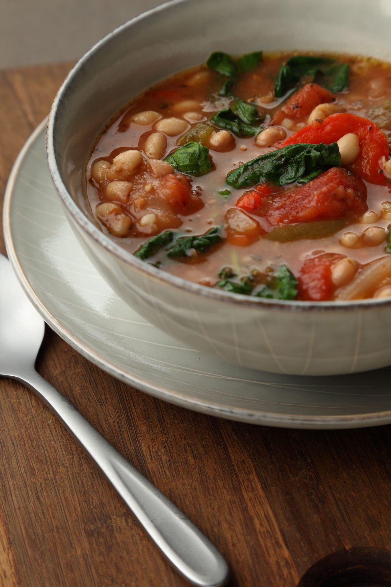 A spoon rests next to saucer that holds a bowl of soup made with vegetables and navy beans.
