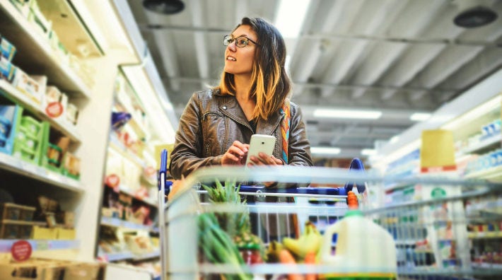 : A woman stands behind a shopping cart that contains milk and produce and looks at a shelf of food at a grocery store. 